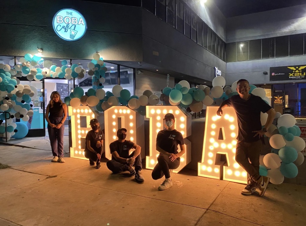 The Boba Café team in front of glowing BOBA marquee letters and balloon arch
